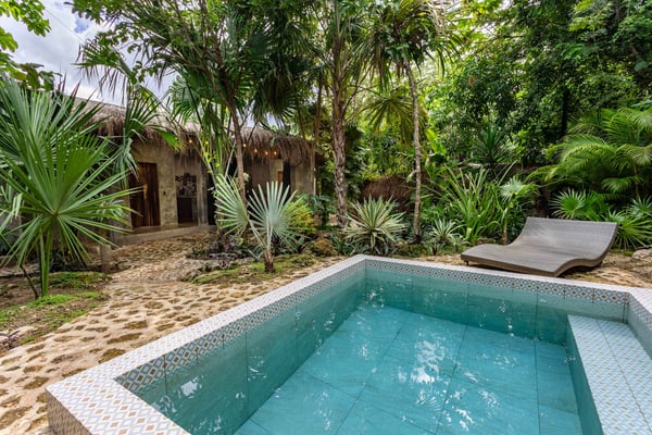 Tropical courtyard with turquoise swimming pool, thatched-roof stone buildings, lounge chair, and dense palm and tree vegetation