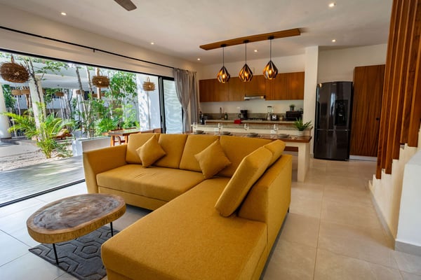 Open-plan living room with mustard yellow sectional sofa, wood coffee table, kitchen with dark wood cabinets, and sliding glass doors to tropical courtyard.
