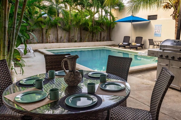 Courtyard with dining table set for four, swimming pool, blue umbrella, lounge chairs, and gas grill surrounded by palm trees