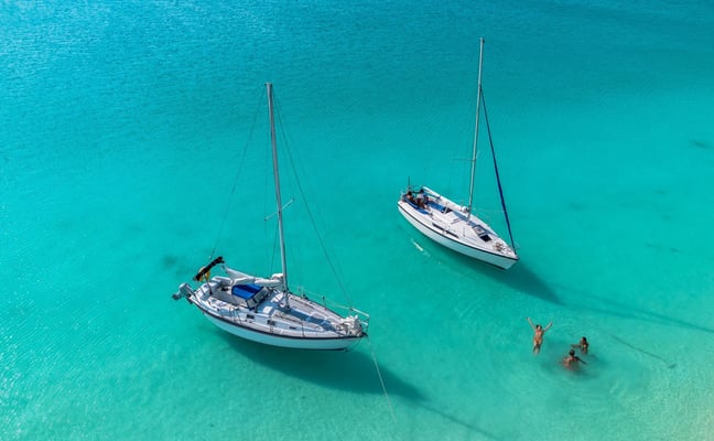 Two sailboats anchored in turquoise water near three people swimming.