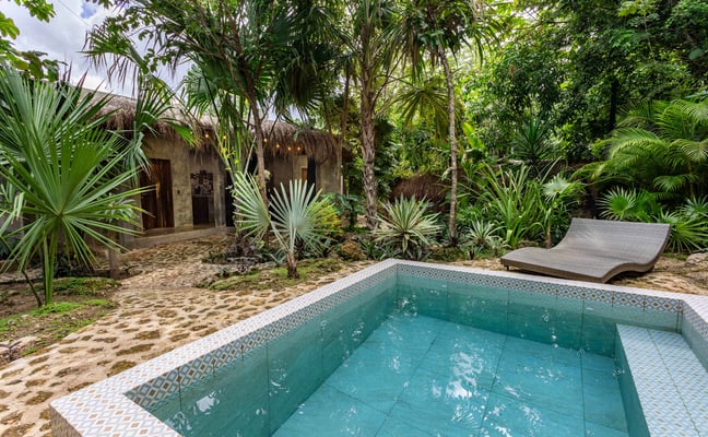 Tropical courtyard with turquoise pool, thatched-roof stone buildings, palm trees, and lounge chair