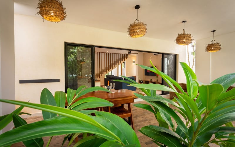Outdoor patio with dining table, greenery, and hanging woven lights. Sliding glass doors lead to interior.