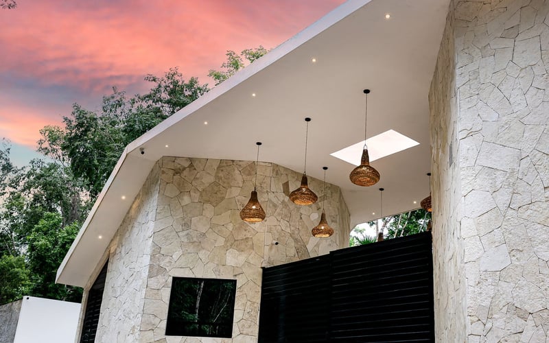 Stone-clad entrance with black gate, pendant lights, under a white roof, against a sunset sky.